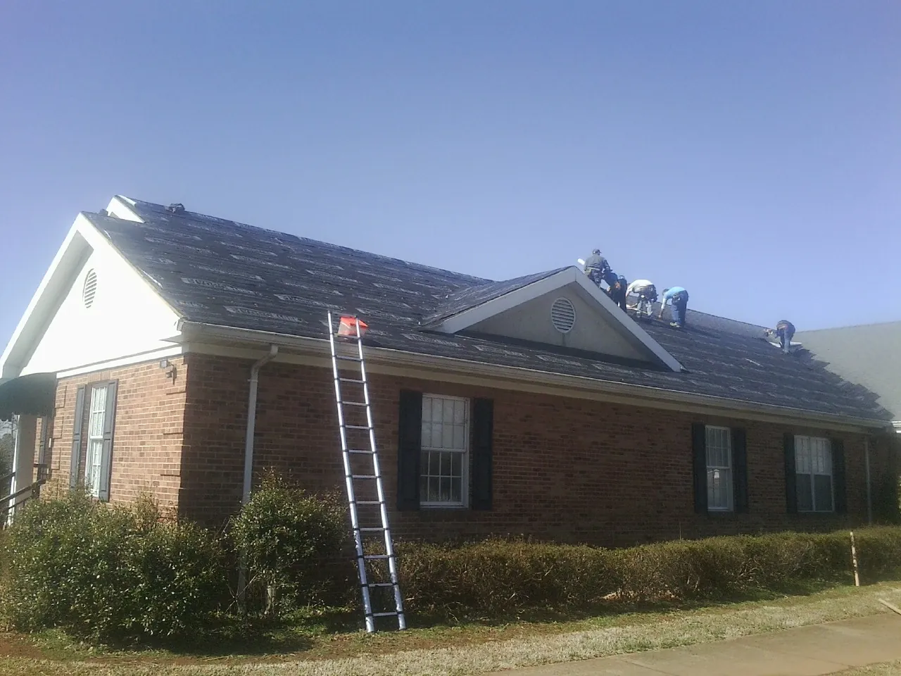 Crew tearing shingles off a brick ranch roof with a ladder set — Emergency Roof Repair work by our crew in Brighton