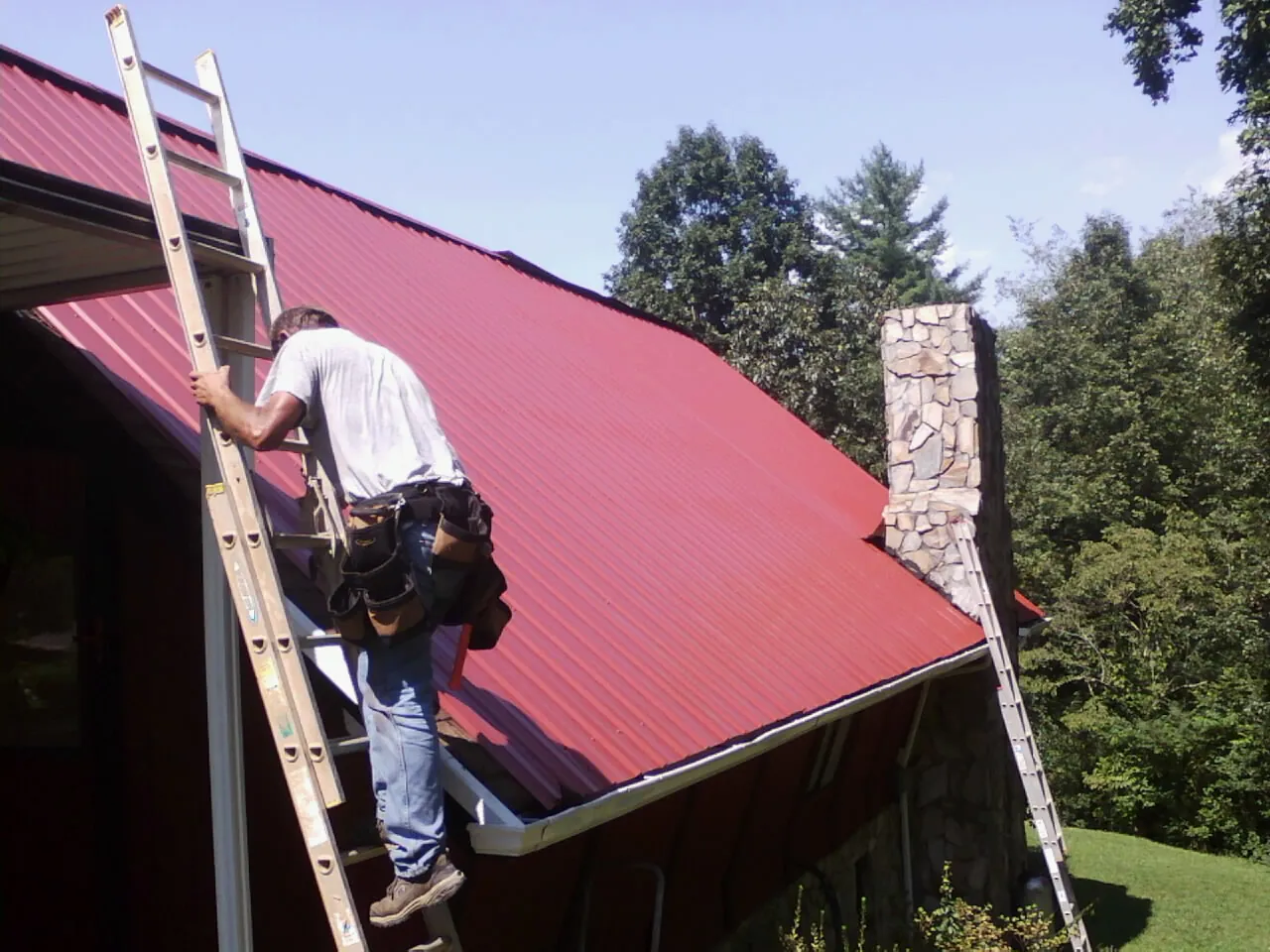 Roofer on a ladder beside a red metal roof and stone chimney — Soffit & Fascia Repair by Roofing Brothers in Brighton